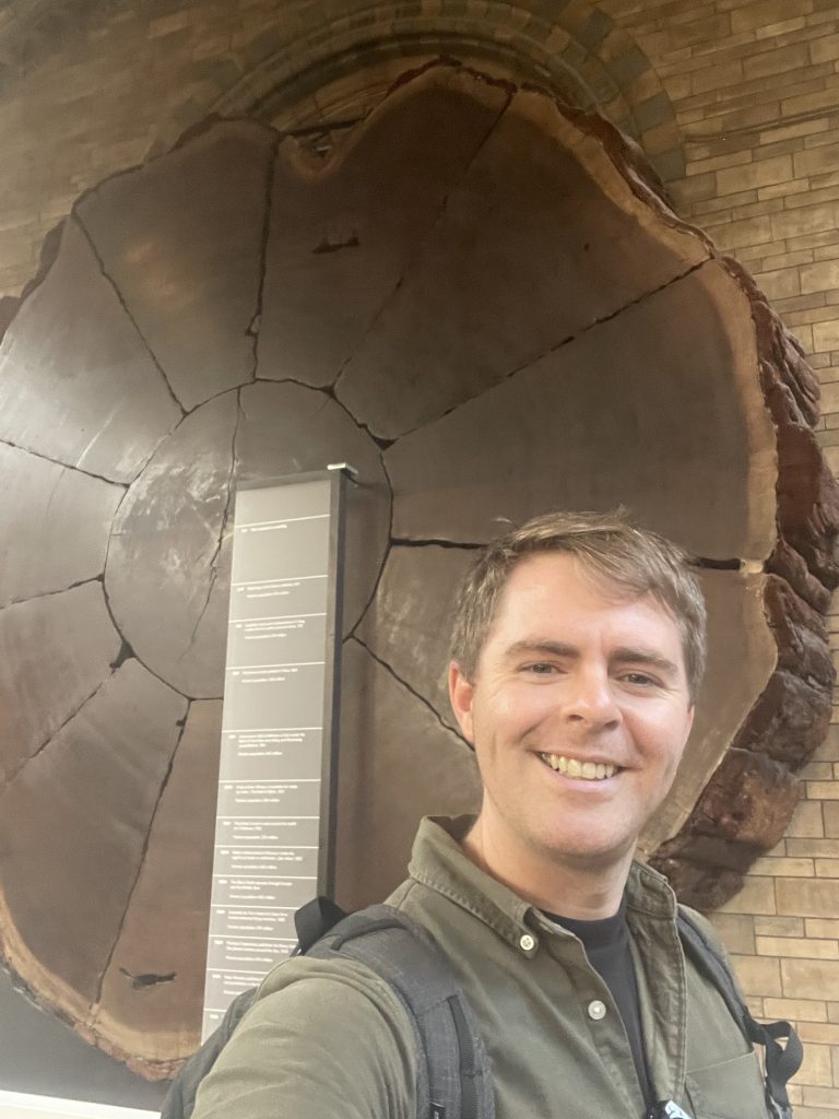 Jason Martin in front of a giant sequoia at the Natural History Museum wearing a khaki shirt.