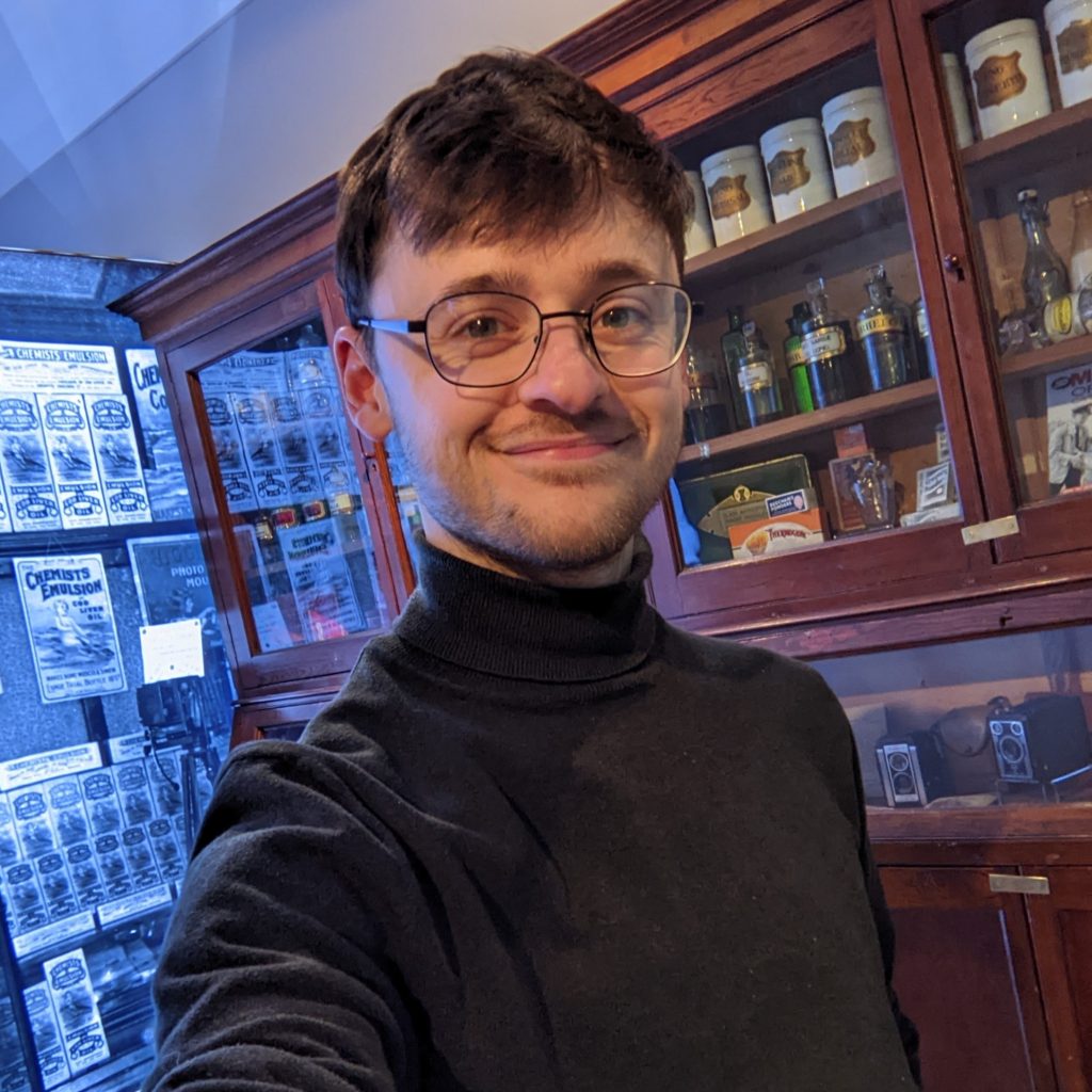 Joe Derry Setch in a black poloneck and glasses in a traditional style pharmacist