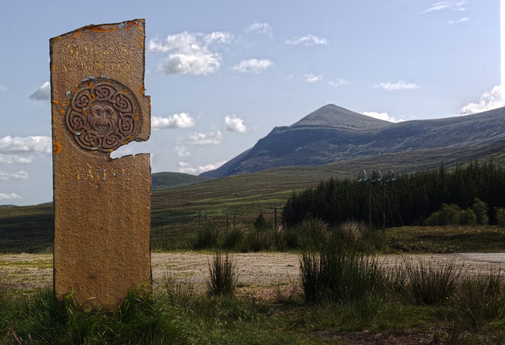 picture of a tall, narrow standing stone with circular image and the word 'failte' written on it. In the background is a a pointed mountain and blue sky with white fluffy clouds