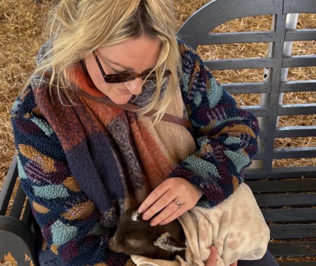 Parris with long blonde hair in a blue and brown cardigan, a brown/orange scarf, wearing sunglasses and holding a brown goat