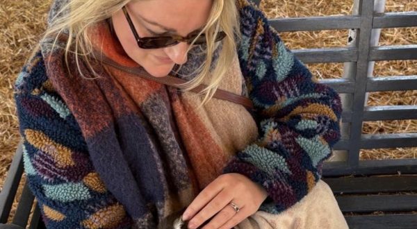 Parris with long blonde hair in a blue and brown cardigan, a brown/orange scarf, wearing sunglasses and holding a brown goat