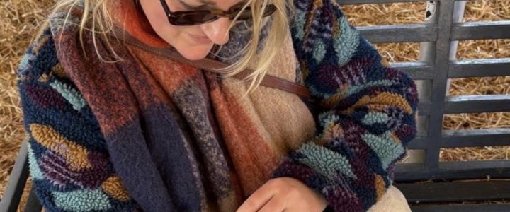 Parris with long blonde hair in a blue and brown cardigan, a brown/orange scarf, wearing sunglasses and holding a brown goat