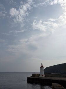White lighthouse standing at the end of a harbour pier, with calm sea, dramatic cliffs and a cloudy Highland sky in the background.
