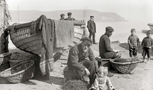 Historic photograph showing people gathered beside fishing boats and baskets on Cromarty shore, with the hills across the water in the background.