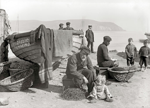 Historic photograph showing people gathered beside fishing boats and baskets on Cromarty shore, with the hills across the water in the background.