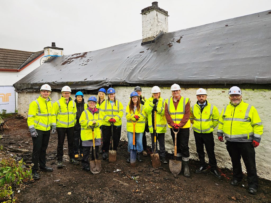 Group of construction workers and project team members wearing high-visibility clothing and hard hats, standing outside a traditional whitewashed stone building with shovels.