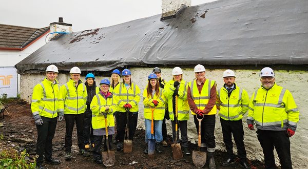 Group of construction workers and project team members wearing high-visibility clothing and hard hats, standing outside a traditional whitewashed stone building with shovels.