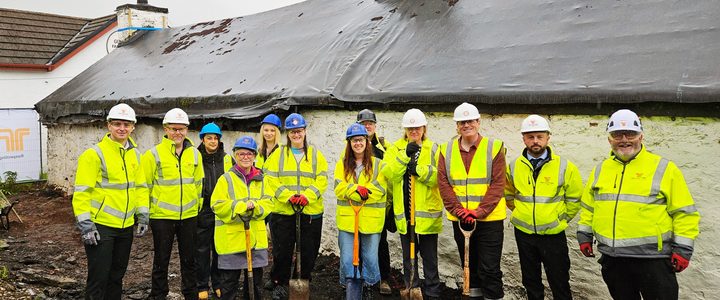 Group of construction workers and project team members wearing high-visibility clothing and hard hats, standing outside a traditional whitewashed stone building with shovels.
