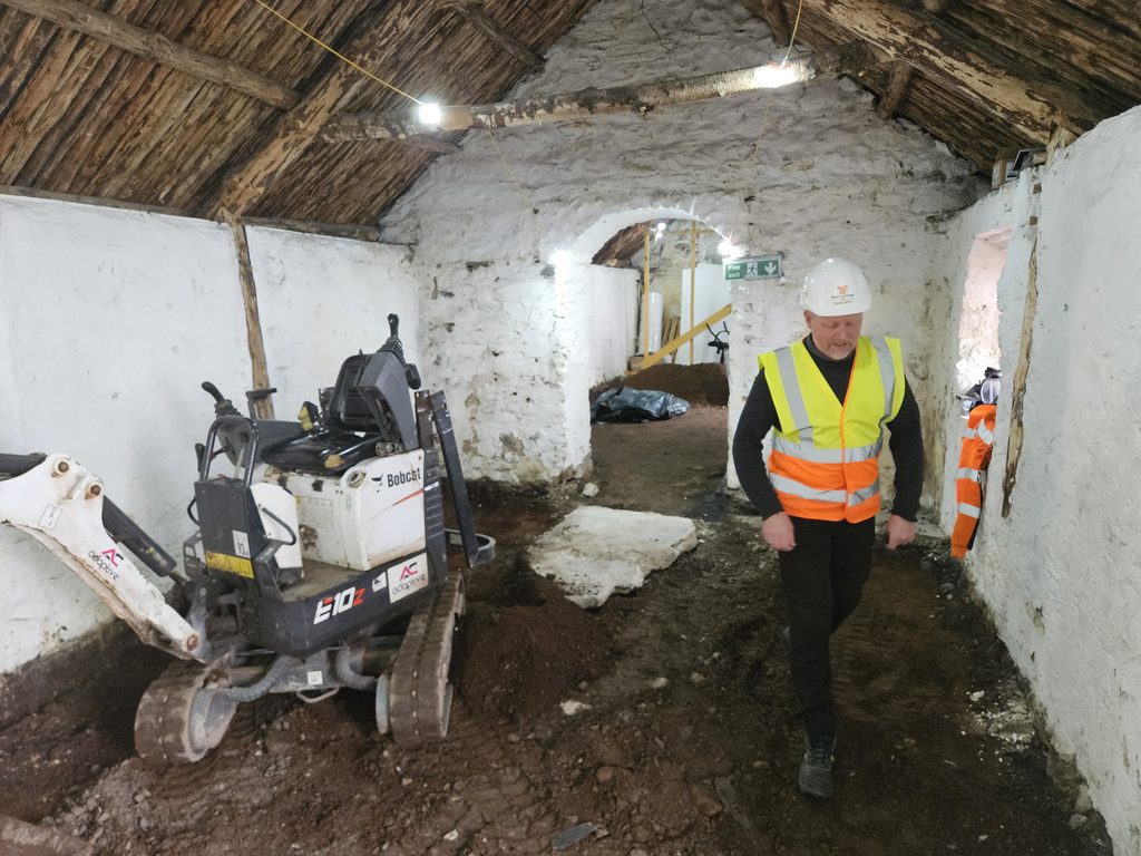 Building works underway inside a traditional stone building, with a small excavator on a partly excavated floor and a worker wearing a hard hat and high-visibility clothing.