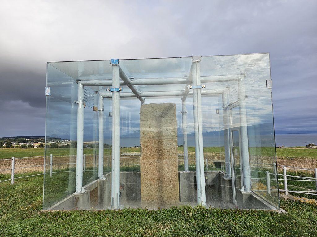 An ornately carved standing stone displayed inside a glass shelter, with open fields and the sea visible in the background.