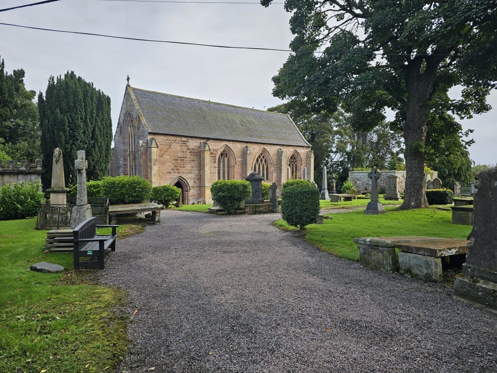A stone church with Gothic arched windows surrounded by a historic graveyard, mature trees, gravestones and green lawns.