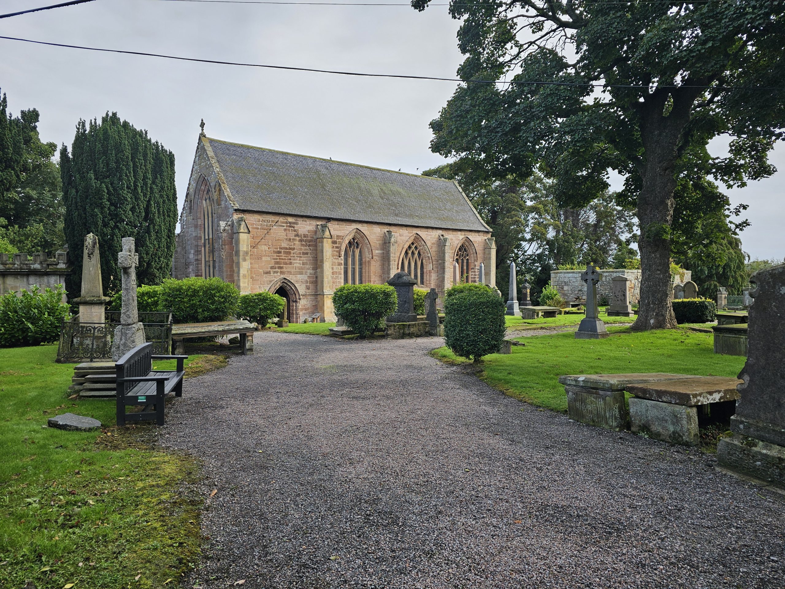 A stone church with Gothic arched windows surrounded by a historic graveyard, mature trees, gravestones and green lawns.